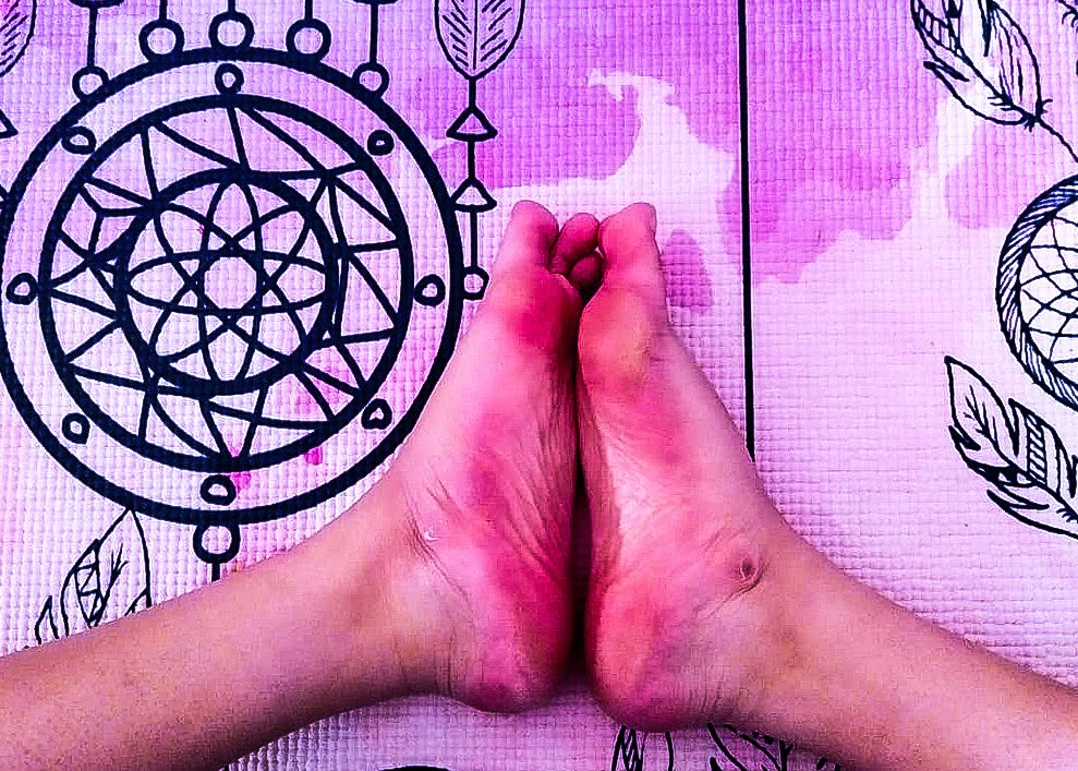 Image shows a close-up of praying feet, with brown skin, on a pink yoga mat with a dreamcatcher motif in black