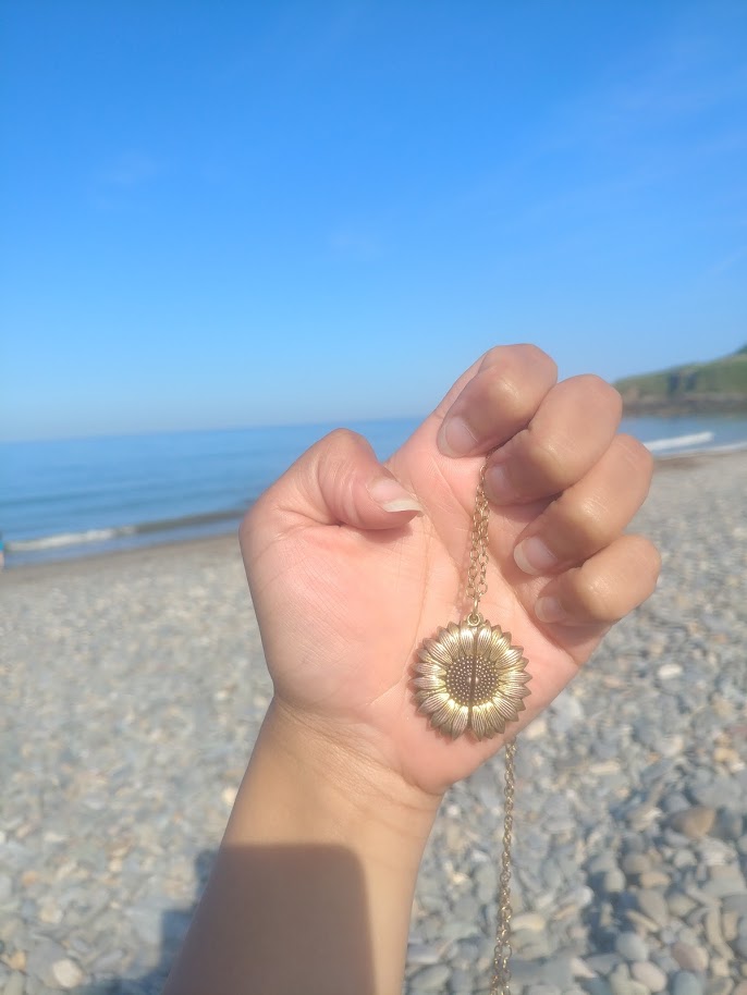 Image shows a hand holding a brass-gold sunflower locket on a chain in the sun. In the background you can see a beach, blue sky and blue sea