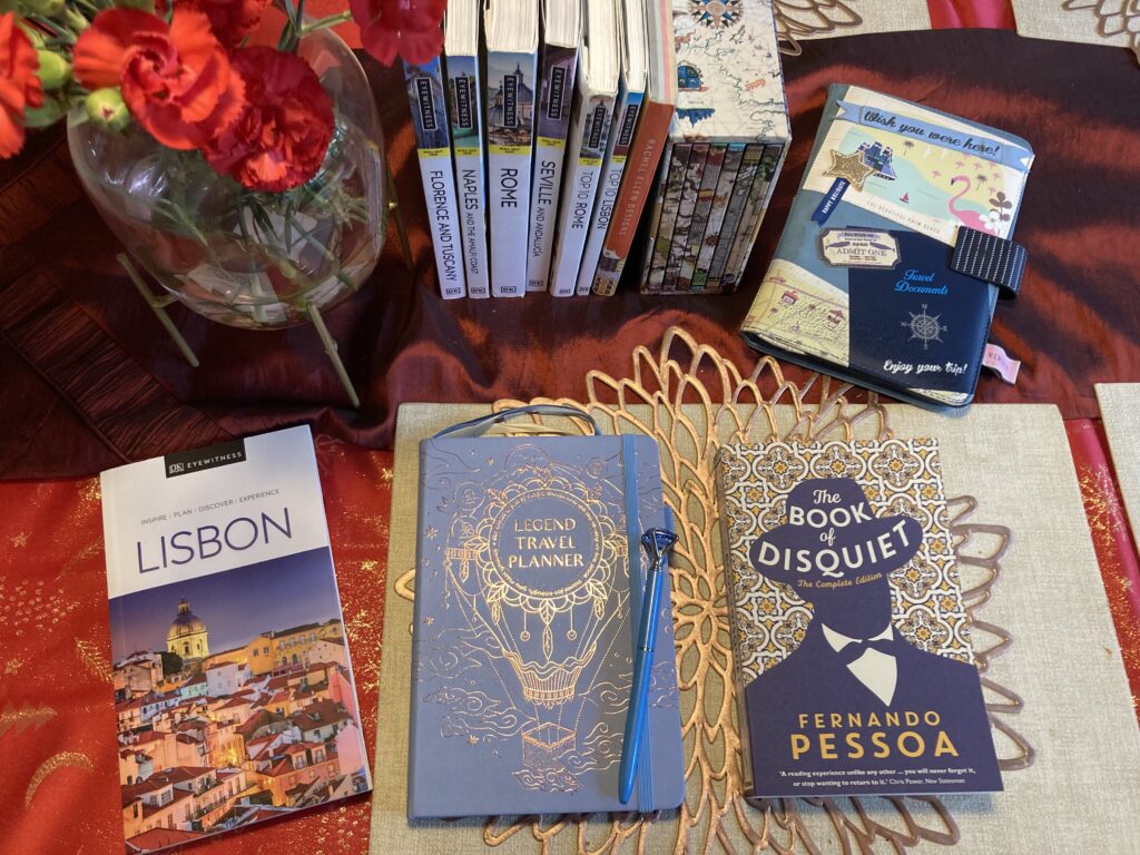 Image shows a collection of travel books and journals on a table with a red tablecloth and some red flowers in the top left corner. On the top half of the page are DK Eyewitness books on Florence, Naples, Rome, Seville and Lisbon. In the top left corner is a colourful travel pouch with flamingos and embellishments. In the bottom half, centre, is a pale blue notebook that says "Legend Travel Planner" in gold, with a light blue pen beside it. The cover in gold depicts a hot air balloon with a dreamcatcher in the centre. To the right, a DK Eyewitness book on Lisbon. To the left, a novel, "The Book of Disquiet", by Fernando Pessoa. 